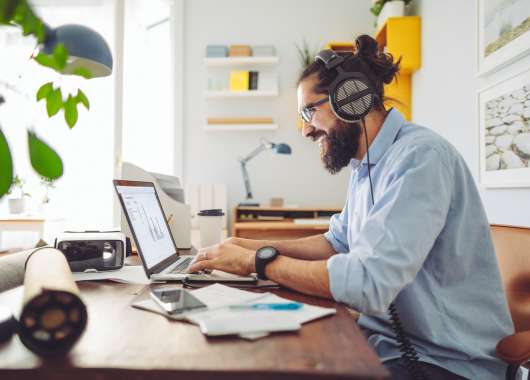 Man wearing headphones and smiling as he types on a laptop in a bright, homey office.