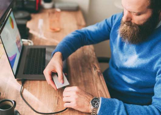 Man in blue sweater with brunette beard plugging external hard drive into laptop.