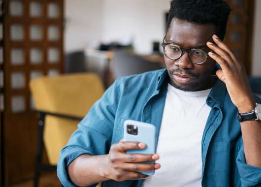 Black man in teal cardigan holding an iPhone with a blue case and looking confused.