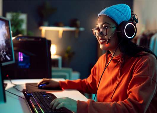 A woman wearing an orange shirt, white beanie with headphones and glasses on uses her computer
