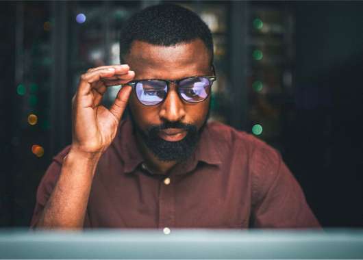 Black man holding his glasses while staring at a computer.