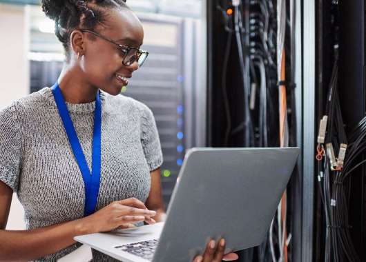 A woman holding a laptop smiles as she looks at a server
