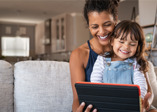 A young child sitting in her mother's lap, smiling and laughing while using a handheld device