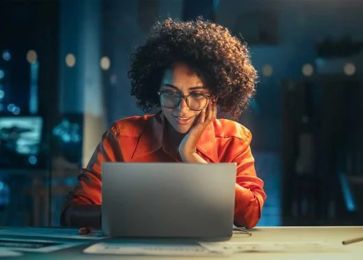 A woman sits at her laptop computer at night, looking intrigued