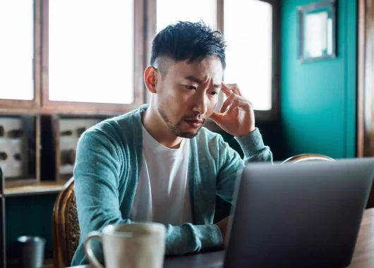A man sits on his desk looking into his laptop screen and appears stressed.